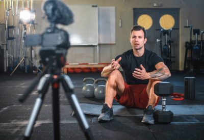 Caucasian male fitness instructor sitting on exercise mat at gym and making vlog explaining how to workout with kettlebells and dumbbells.
